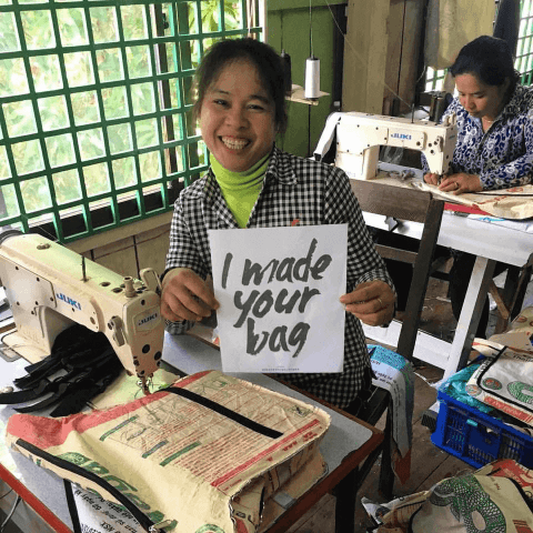 A woman sits next to a sewing machine holding a sign that says 'I made your bag'